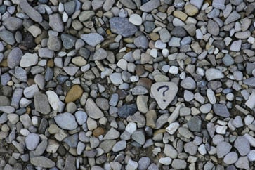 A collection of small, multicolored pebbles with one larger stone in the center marked with a hand-drawn question mark, standing out among the scattered rocks.