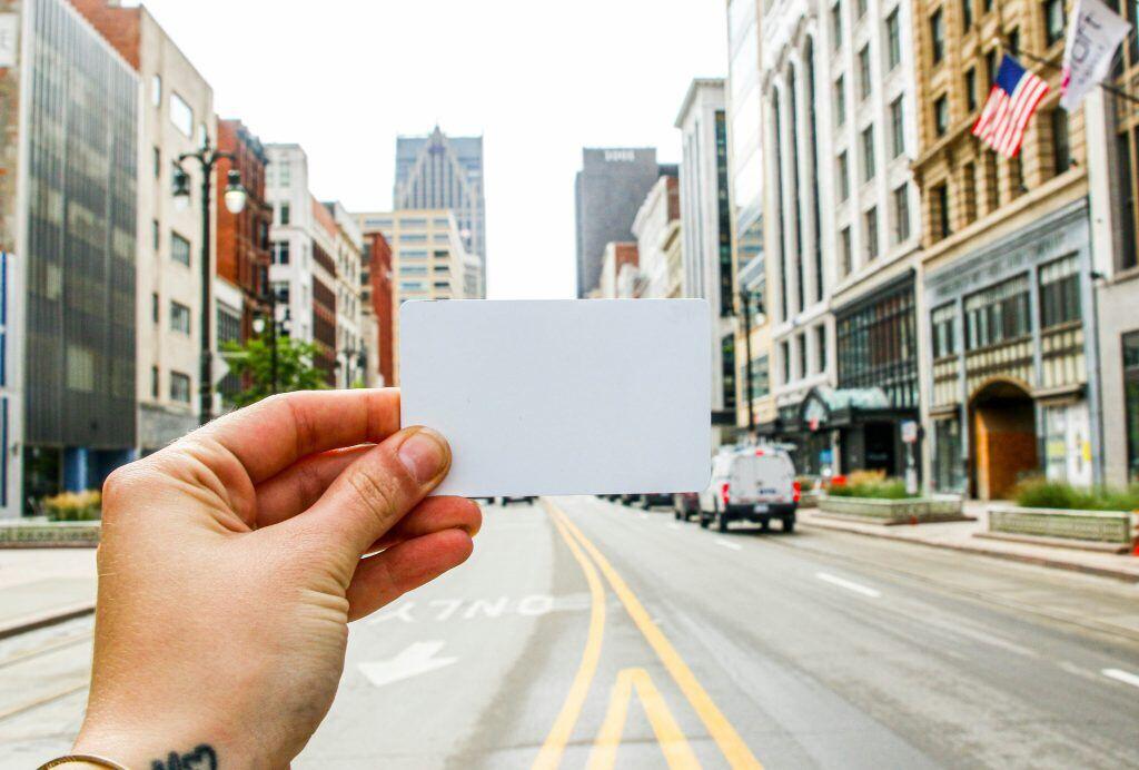 Hand holding a blank card in the foreground on a city street, with buildings and an American flag visible in the background.