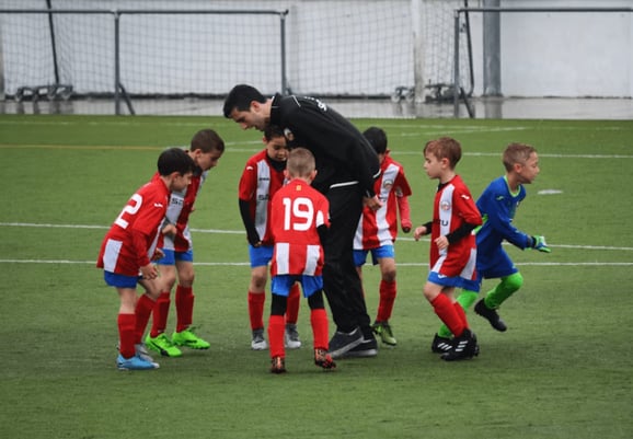 Coach guiding young soccer players in red and white uniforms during practice on a green field, emphasizing teamwork and preparation.