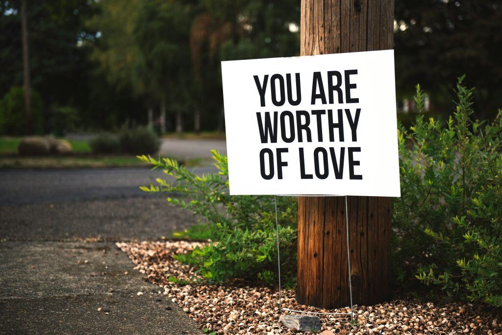 Sign reading 'You Are Worthy of Love' placed in front of a wooden post along a roadside.